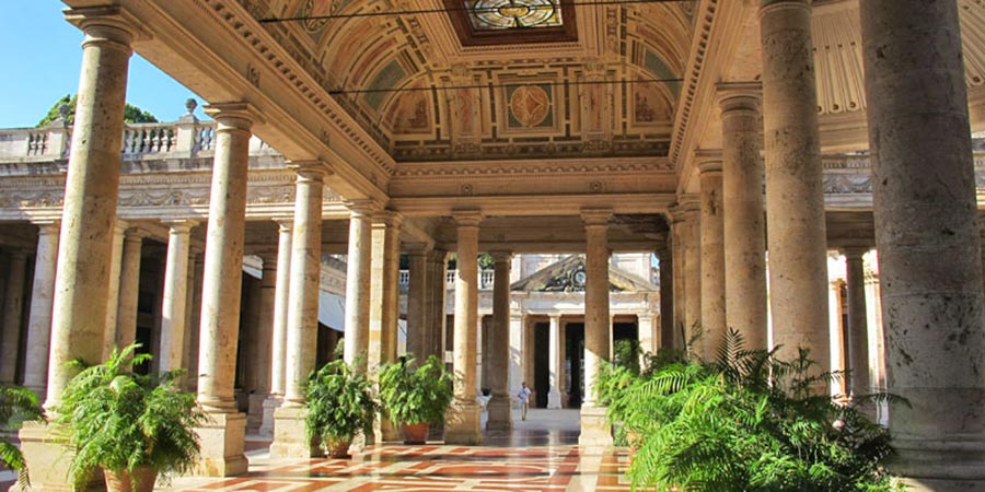 Exterior view of the Grand Hotel Francia & Quirinale, a luxurious, historic hotel. The grand building features elegant architecture with classical columns with a view down an exterior corridor lined with lush green planters, with the ceiling encompassing a stained-glass sky light and curved painted details. 